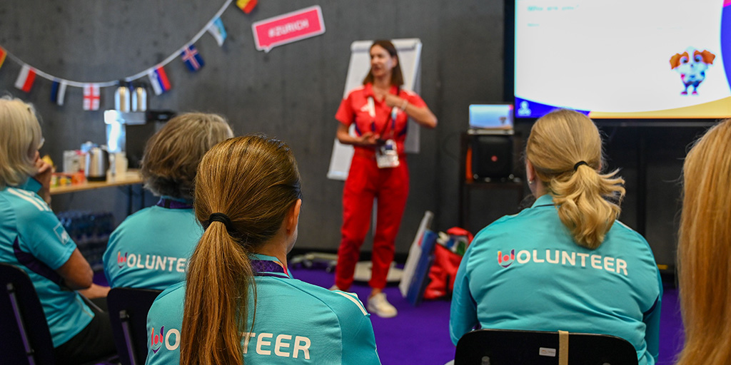 A group of volunteers at a briefing, with their backs to the camera, while an instructor addresses them from the front of the room